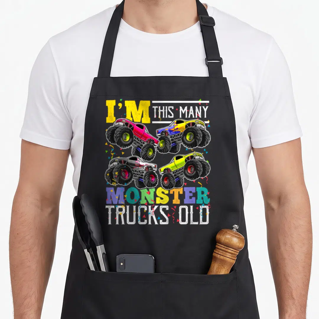 Many truck apron - Four monster trucks and confetti with I'M THIS MANY MONSTER TRUCKS OLD text — shown on a black cotton canvas apron against a white background