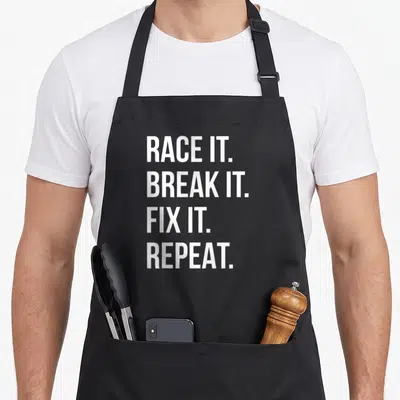 Race drag apron - White stacked text reading RACE IT. BREAK IT. FIX IT. REPEAT — shown on a black cotton canvas apron against a white background