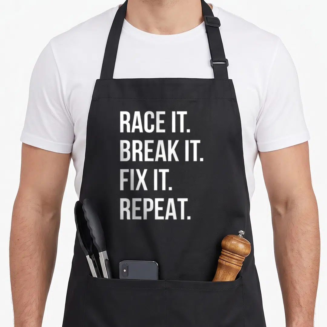 Race drag apron - White stacked text reading RACE IT. BREAK IT. FIX IT. REPEAT — shown on a black cotton canvas apron against a white background