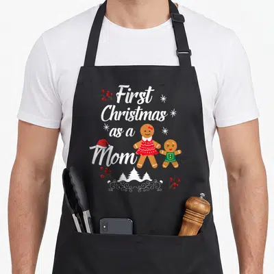 Xmas christmas aprons - First Christmas as a Mom text with two gingerbread people and holly berries — shown on a black cotton canvas apron against a white background
