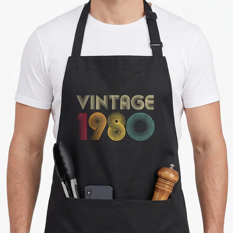 46th birthday apron - Vintage 1980 text in beige red yellow teal retro stripes — shown on a black cotton canvas apron against a white background