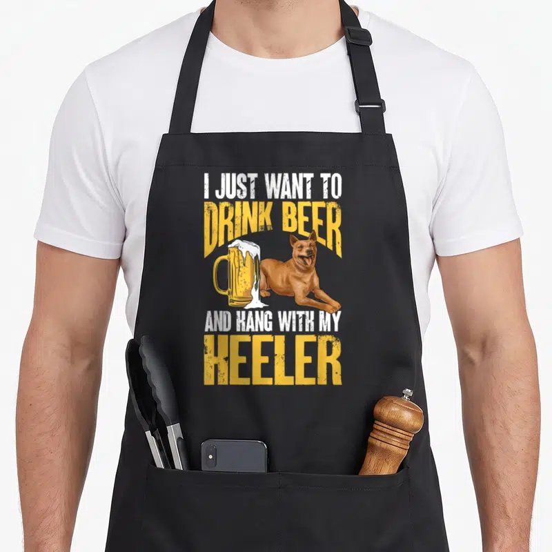 Heeler drinking dog apron - Lying brown dog next to overflowing beer mug with distressed yellow and white text — shown on a black cotton canvas apron against a white background