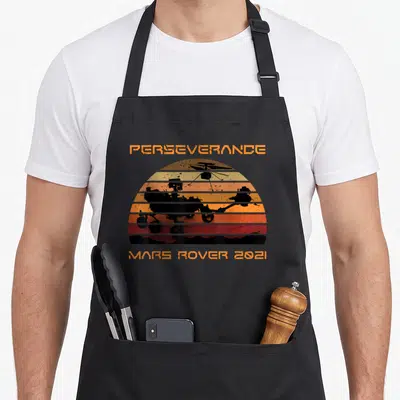 Perseverance moon landing apron - Retro orange red sun with black Mars rover and helicopter silhouettes — shown on a black cotton canvas apron against a white background