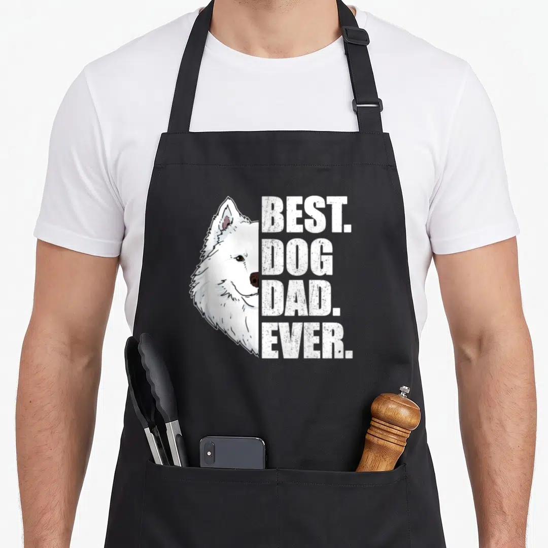 Samoyed father day apron - White cartoon Samoyed head beside stacked BEST DOG DAD EVER text in black outline — shown on a black cotton canvas apron against a white background