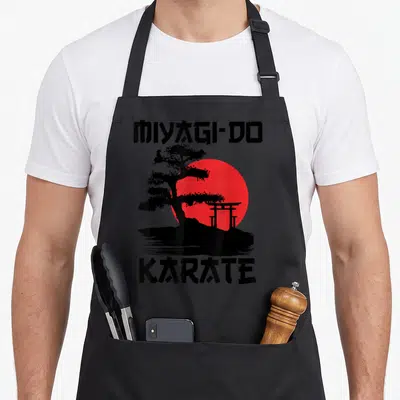 Miyagi road apron - Bonsai tree and torii gate silhouette against a red sun with black text reading MIYAGI-DO and KARATE — shown on a black cotton canvas apron against a white background