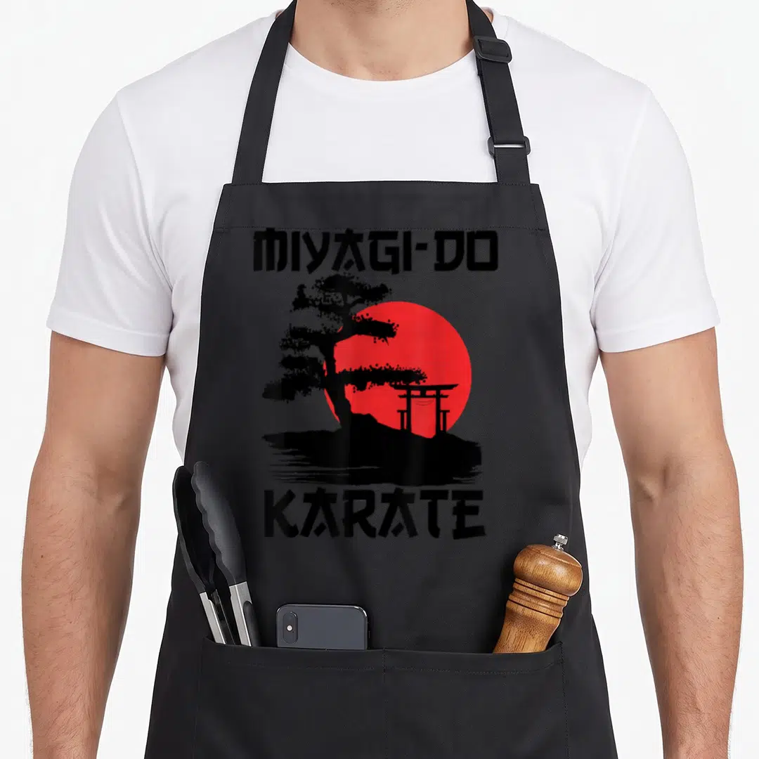 Miyagi road apron - Bonsai tree and torii gate silhouette against a red sun with black text reading MIYAGI-DO and KARATE — shown on a black cotton canvas apron against a white background