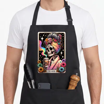 Tarot messy aprons - Skeleton in turban holding coffee cup with THE MOM text and floral border — shown on a black cotton canvas apron against a white background