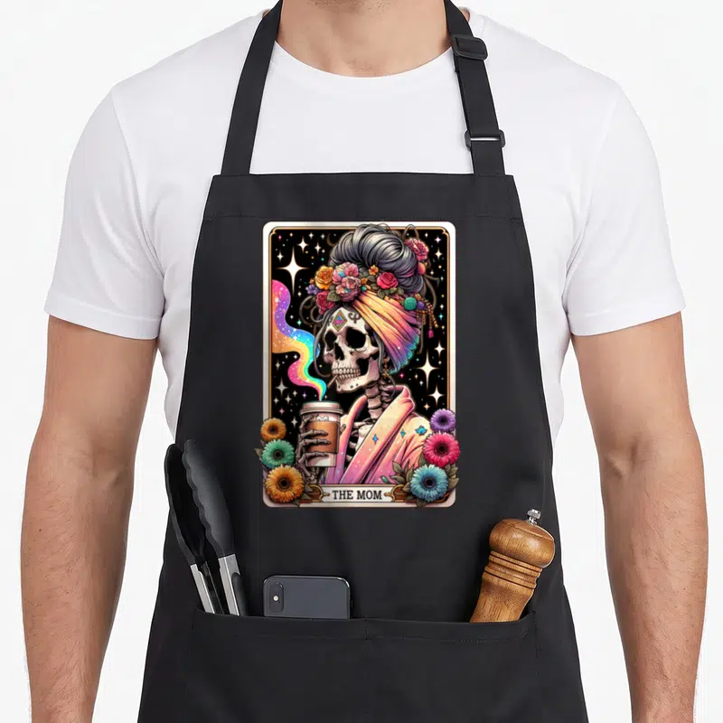 Tarot messy aprons - Skeleton in turban holding coffee cup with THE MOM text and floral border — shown on a black cotton canvas apron against a white background