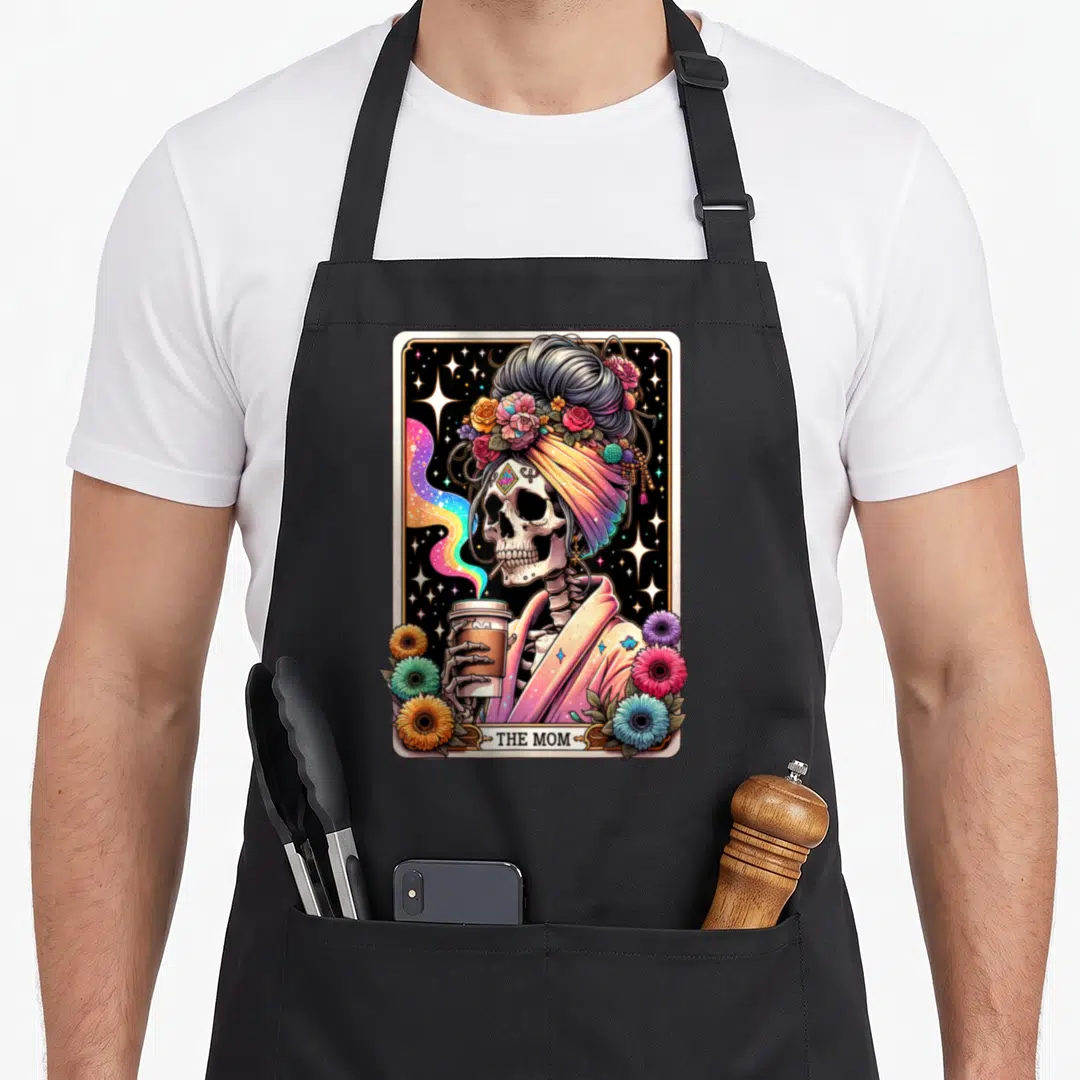 Tarot messy aprons - Skeleton in turban holding coffee cup with THE MOM text and floral border — shown on a black cotton canvas apron against a white background