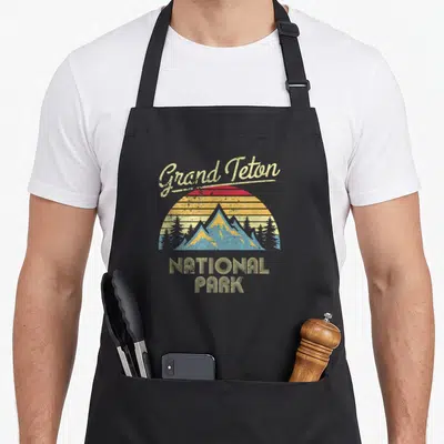 Funny grand teton national park apron - Retro Grand Teton mountain graphic with arched script and striped sun in beige — shown on a black cotton canvas apron against a white background