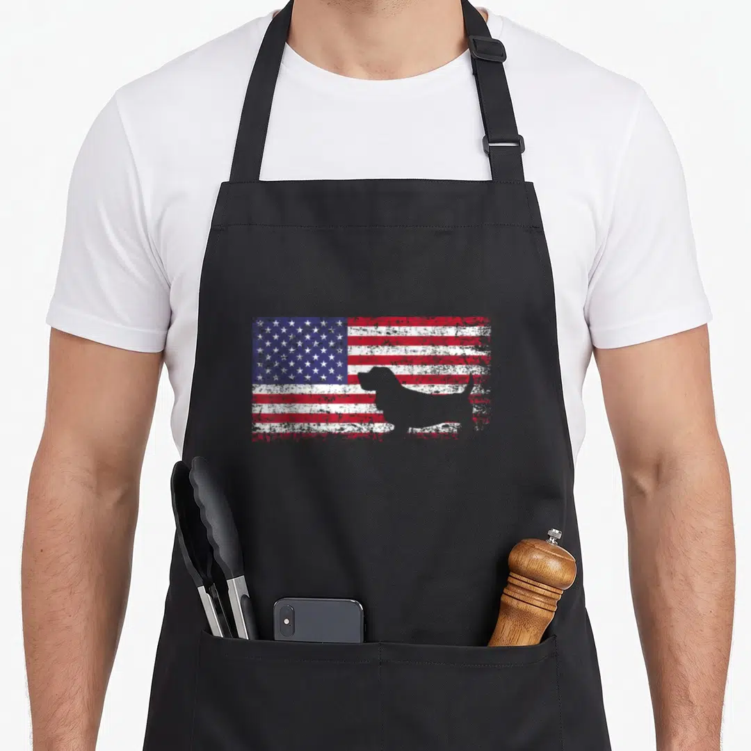 Basset american flag apron - Grey basset hound silhouette on distressed red white and blue flag — shown on a black cotton canvas apron against a white background