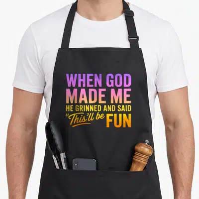 Grinned novelty apron - Stacked colorful text reading When God Made Me He Grinned and Said This'll Be Fun — shown on a black cotton canvas apron against a white background