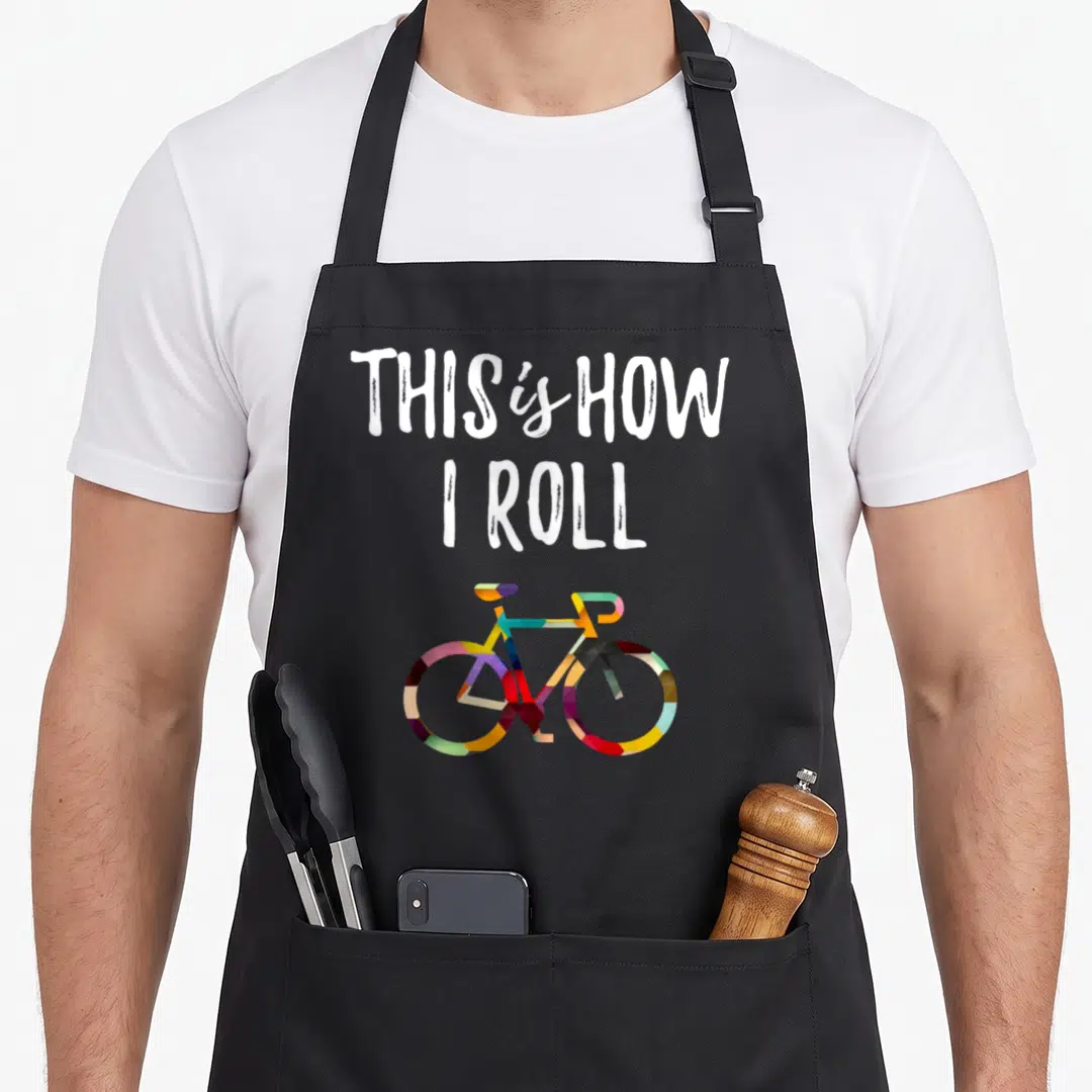 Bicycle colorful aprons - Stacked white text THIS is HOW I ROLL above a multicolored geometric bicycle — shown on a black cotton canvas apron against a white background
