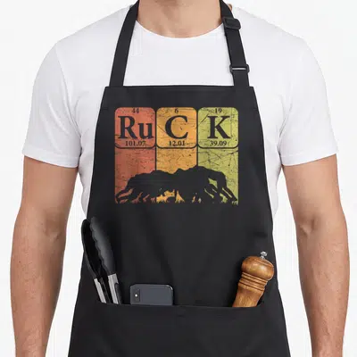 Ruck table apron - Periodic table cells Ru C K with rugby silhouette on red orange yellow distressed background — shown on a black cotton canvas apron against a white background