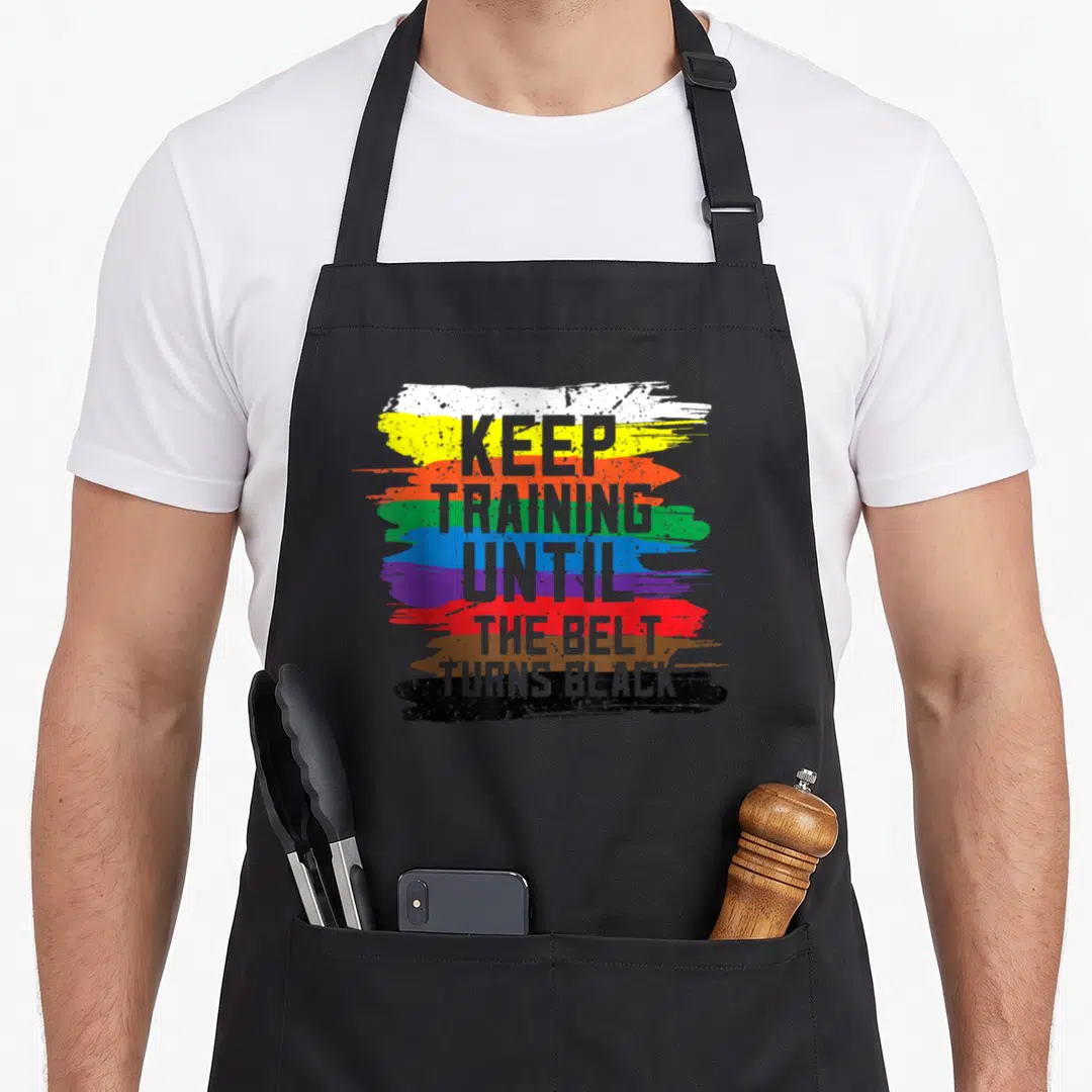 Martial training apron - Colorful distressed brush strokes with grey text reading KEEP TRAINING UNTIL THE BELT TURNS — shown on a black cotton canvas apron against a white background