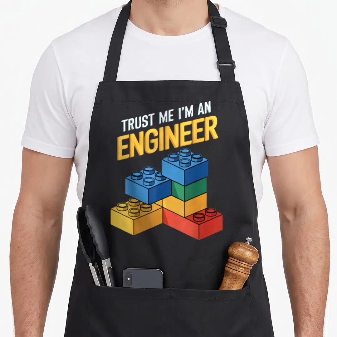 Engineer brick block & apron - Stacked colorful building blocks under arched text reading TRUST ME I'M AN ENGINEER — shown on a black cotton canvas apron against a white background