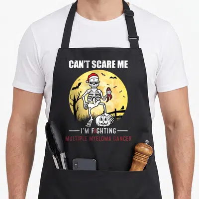 Multiple halloween apron - Skeleton holding red ribbon in front of yellow moon with Can't Scare Me text — shown on a black cotton canvas apron against a white background