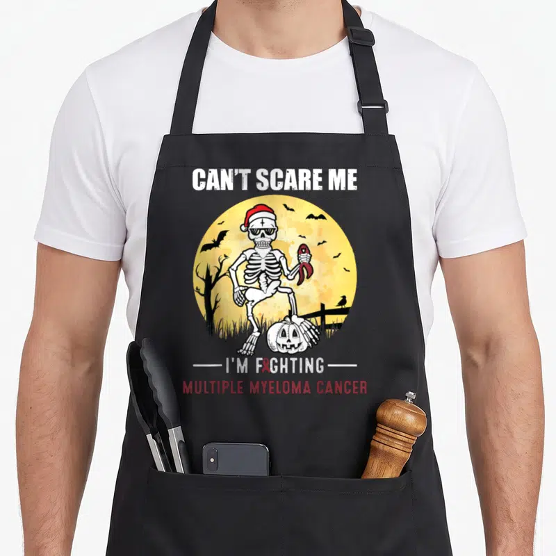 Multiple halloween apron - Skeleton holding red ribbon in front of yellow moon with Can't Scare Me text — shown on a black cotton canvas apron against a white background