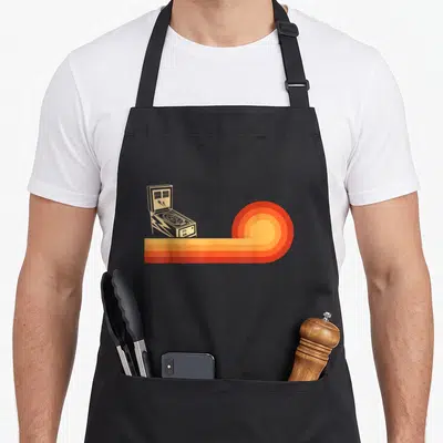 Funny pinball arcade apron - Beige pinball machine with red orange yellow concentric stripes curling to the right — shown on a black cotton canvas apron against a white background