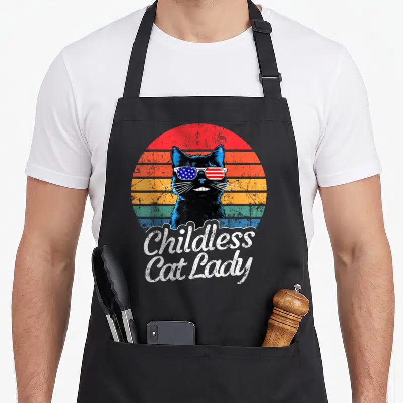 Childless voting cat lady apron - Black cat with flag sunglasses and striped circle below Childless Cat Lady text — shown on a black cotton canvas apron against a white background