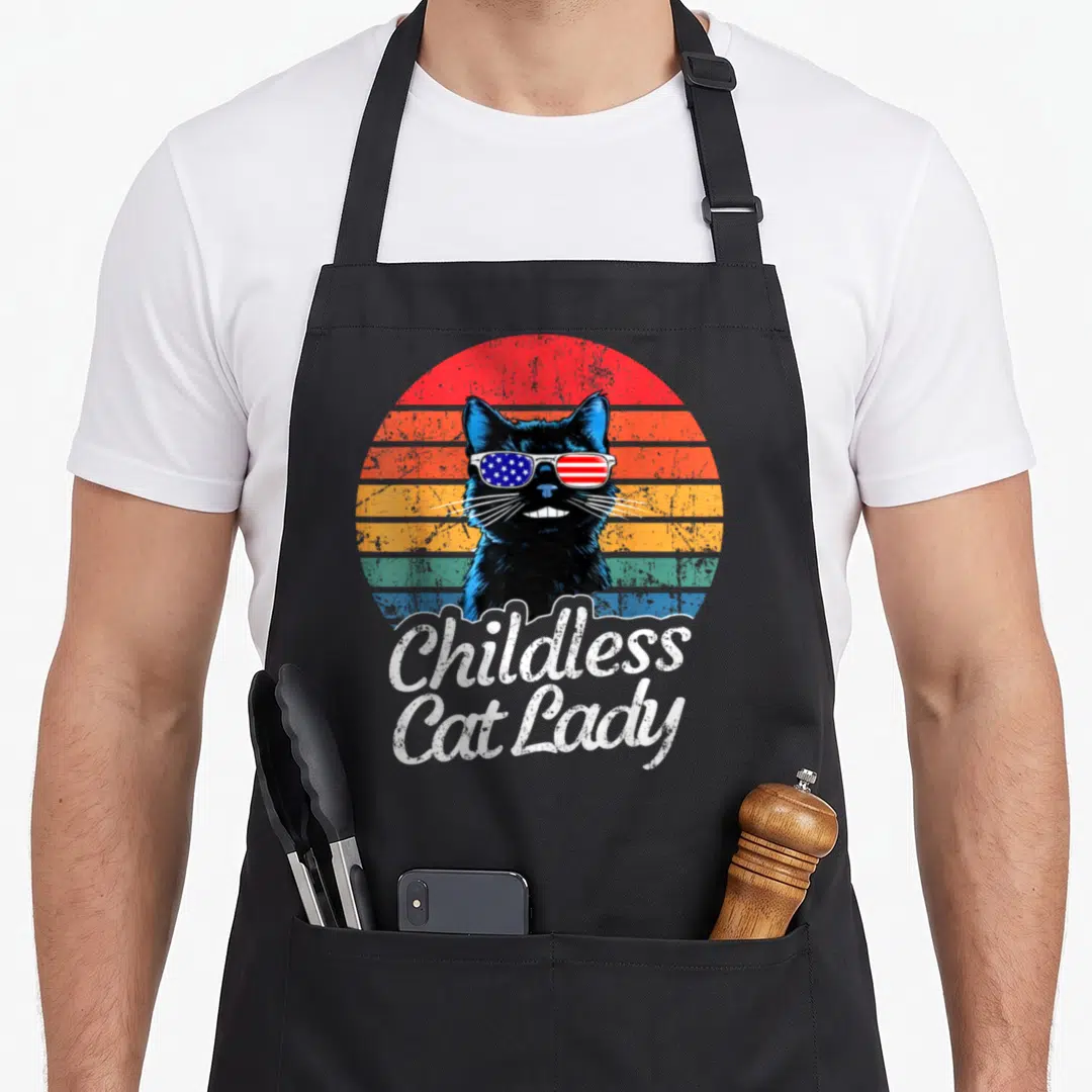 Childless voting cat lady apron - Black cat with flag sunglasses and striped circle below Childless Cat Lady text — shown on a black cotton canvas apron against a white background