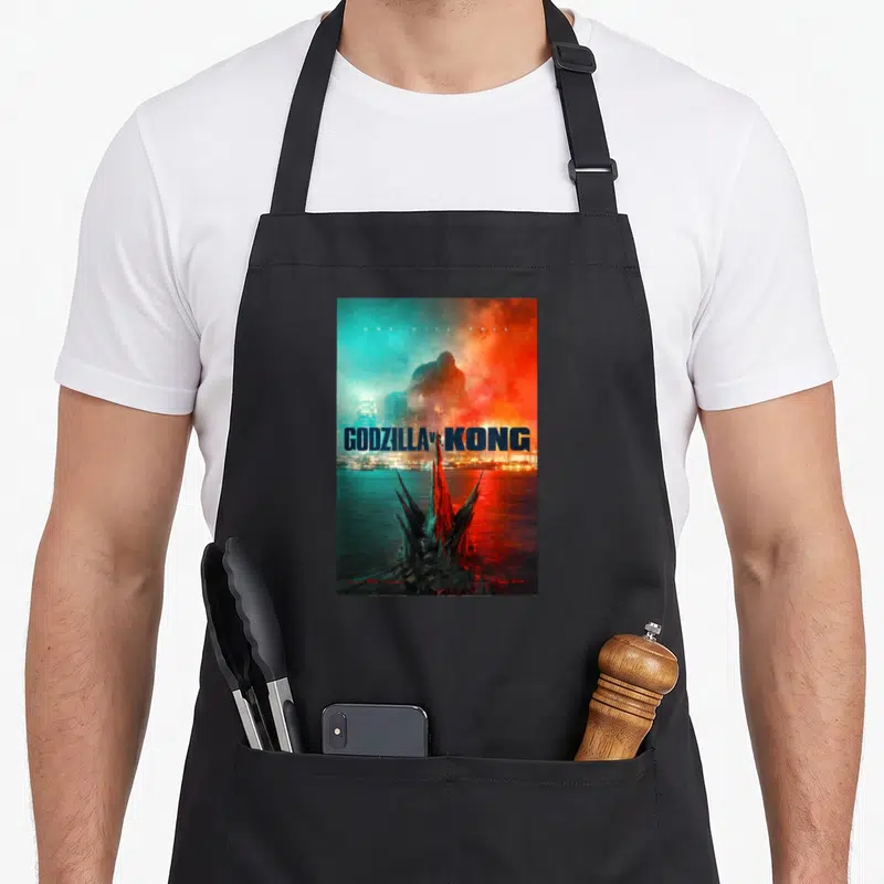 Monsterverse honestly apron - Godzilla fins in teal water with Kong silhouette above city and movie title text — shown on a black cotton canvas apron against a white background