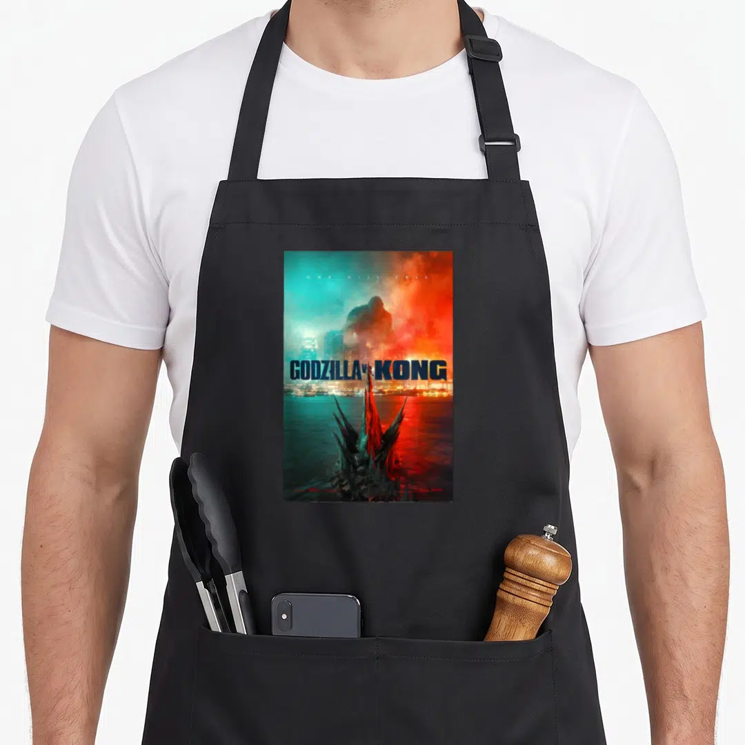 Monsterverse honestly apron - Godzilla fins in teal water with Kong silhouette above city and movie title text — shown on a black cotton canvas apron against a white background