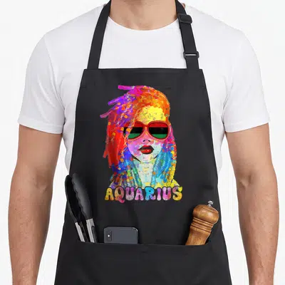 Zodiac hair apron - Watercolor woman with dreadlocks and red sunglasses, AQUARIUS text below in multicolor letters — shown on a black cotton canvas apron against a white background