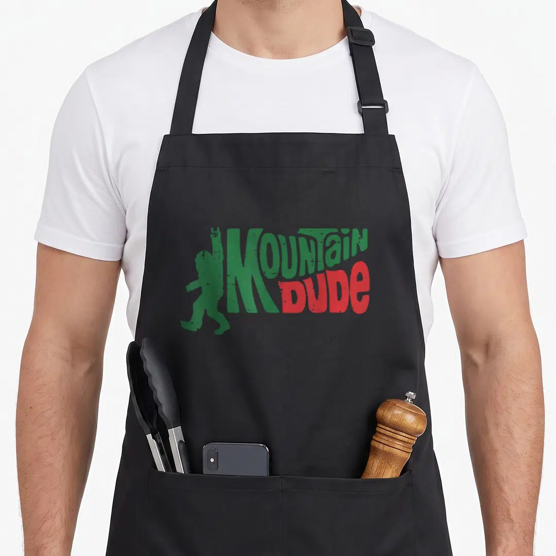 Hiking dude apron - Green Bigfoot silhouette raising hand next to distressed MOUNTAIN DUDE text in green and red — shown on a black cotton canvas apron against a white background