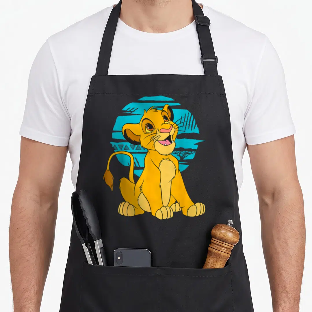 Young lion apron - Yellow cartoon lion cub sitting in front of blue circle with geometric patterns — shown on a black cotton canvas apron against a white background