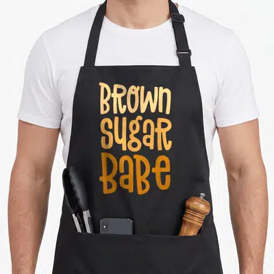 Sugar pride apron - Stacked brown sugar babe text in light to dark orange gradient typography — shown on a black cotton canvas apron against a white background