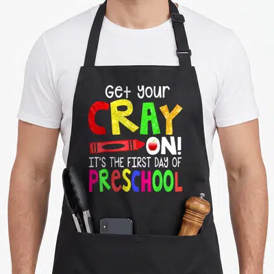 Crayon preschool apron - Glittery text Get your CRAY ON with red crayon and apple illustration — shown on a black cotton canvas apron against a white background