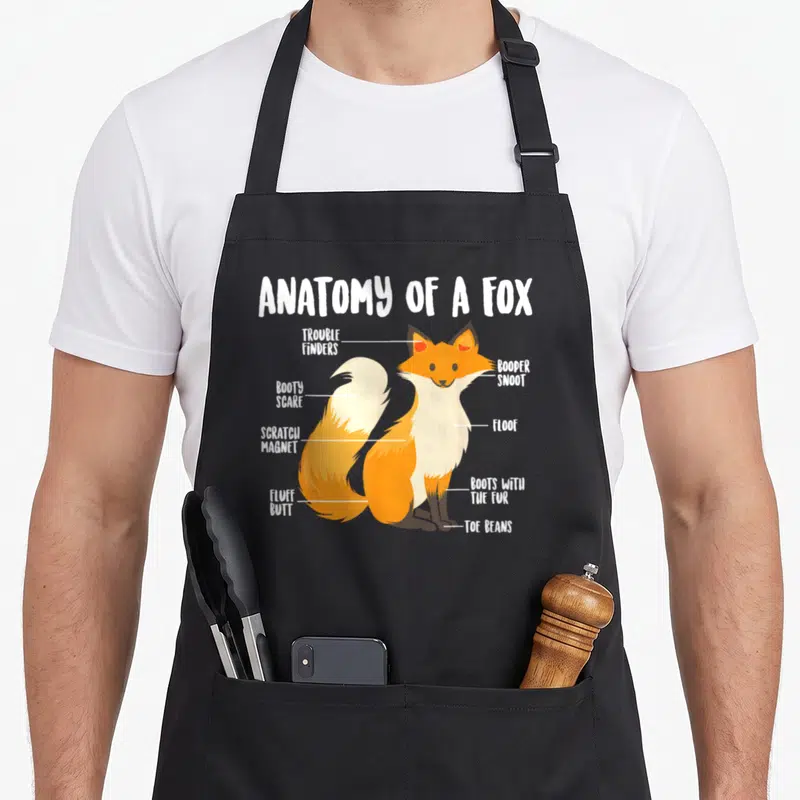 Fox anatomy apron - Cartoon fox diagram labeled with funny body part names like Booty Scare and Toe Beans — shown on a black cotton canvas apron against a white background