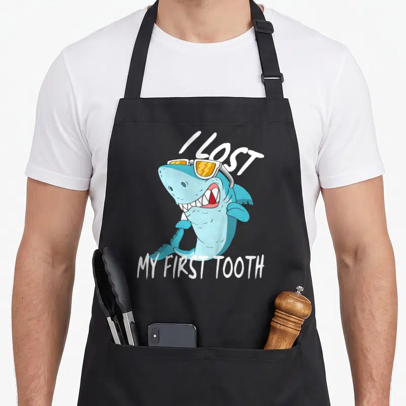 Fairy first tooth apron - Cartoon shark with sunglasses holding a tooth under I lost my first tooth text — shown on a black cotton canvas apron against a white background