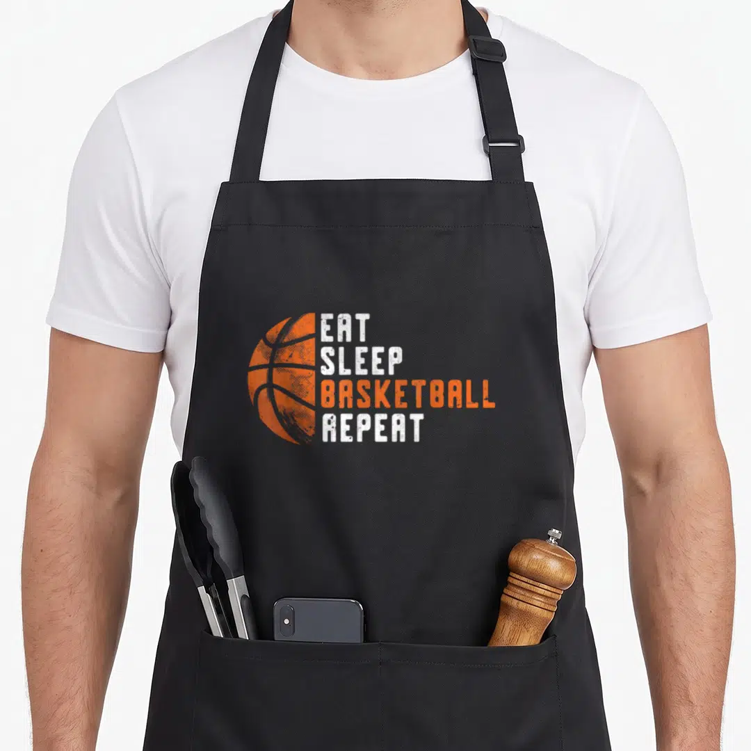 Coach basketball back apron - Distressed orange basketball next to stacked EAT SLEEP BASKETBALL REPEAT text — shown on a black cotton canvas apron against a white background