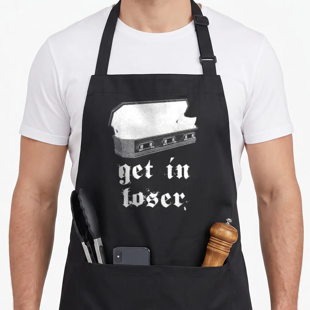 Edgy gothic apron - White distressed open coffin illustration above stacked gothic text reading get in loser — shown on a black cotton canvas apron against a white background