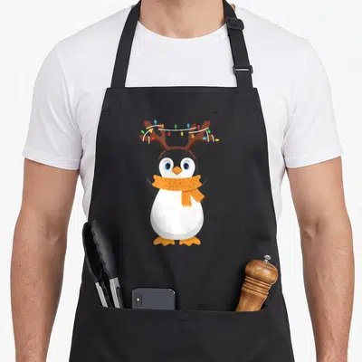 Penguin reindeer apron - Cartoon penguin with brown antlers, wrapped lights, and orange scarf — shown on a black cotton canvas apron against a white background