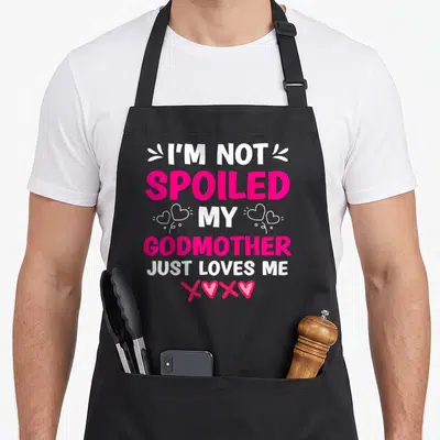 Spoiled matching thanksgiving apron - Stacked text I'm Not Spoiled My Godmother Just Loves Me with white and pink — shown on a black cotton canvas apron against a white background