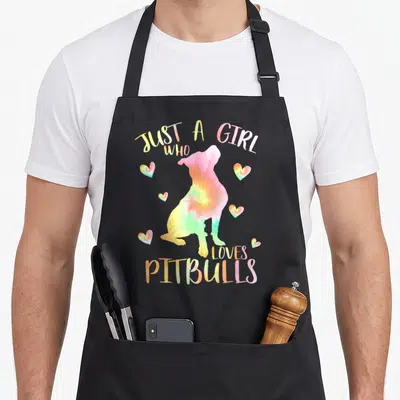 Pitbull tie-dye apron - Tie-dye pitbull silhouette surrounded by hearts and arched text reading Just a Girl Who Loves — shown on a black cotton canvas apron against a white background
