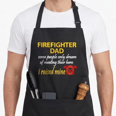 Funny son firefighter aprons - Yellow and white text reading Firefighter Dad next to a red emblem with crossed axes — shown on a black cotton canvas apron against a white background