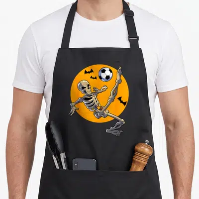 Soccer skeleton player halloween apron - Cartoon skeleton kicking soccer ball on orange circle with three black bats — shown on a black cotton canvas apron against a white background