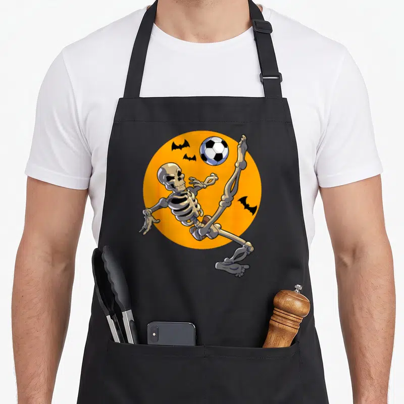 Soccer skeleton player halloween apron - Cartoon skeleton kicking soccer ball on orange circle with three black bats — shown on a black cotton canvas apron against a white background