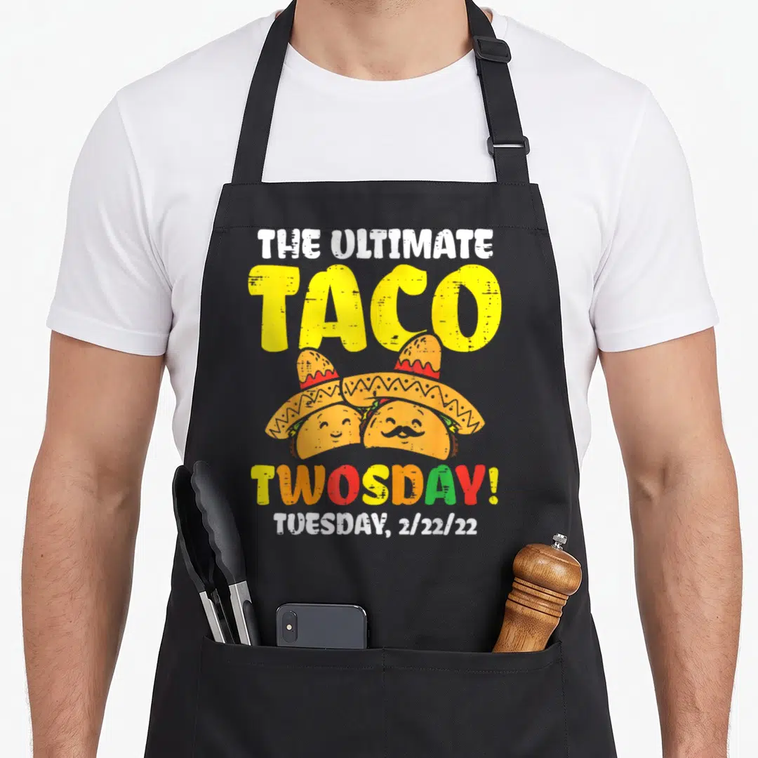 Taco ultimate apron - Two cartoon tacos wearing sombreros under Ultimate Taco Twosday text — shown on a black cotton canvas apron against a white background