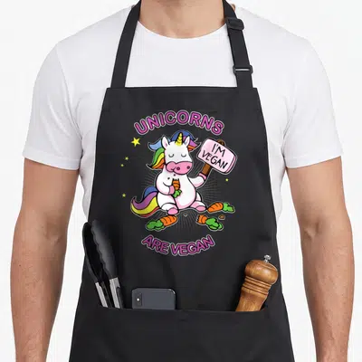 Unicorn veggie apron - Cartoon unicorn holding I'M VEGAN sign and carrot with scattered vegetables and stars — shown on a black cotton canvas apron against a white background