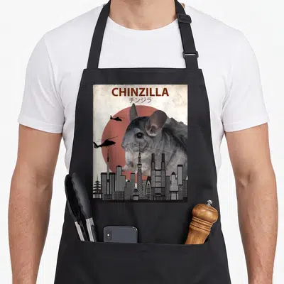 Chinzilla chinchilla lover apron - Giant grey chinchilla behind black skyline and red sun with brown text — shown on a black cotton canvas apron against a white background