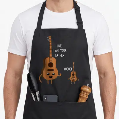Uke playing ukulele apron - Cartoon brown guitar and small uke with white text saying Uke I Am Your Father — shown on a black cotton canvas apron against a white background