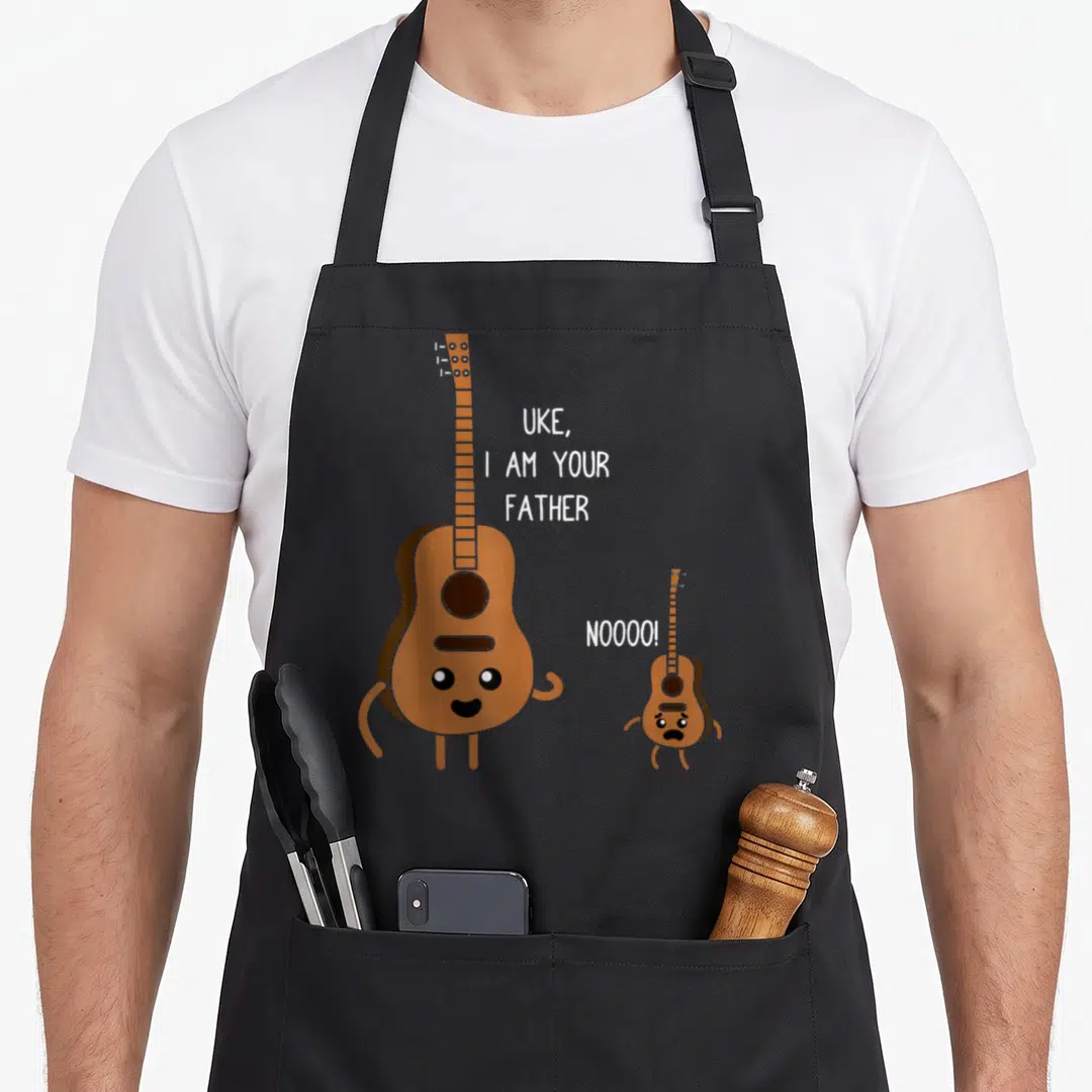 Uke playing ukulele apron - Cartoon brown guitar and small uke with white text saying Uke I Am Your Father — shown on a black cotton canvas apron against a white background