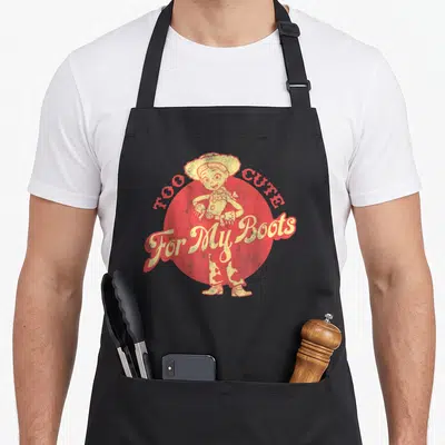 Jessie camping apron - Cartoon girl in cowgirl gear standing in a red circle with text Too Cute For My Boots — shown on a black cotton canvas apron against a white background