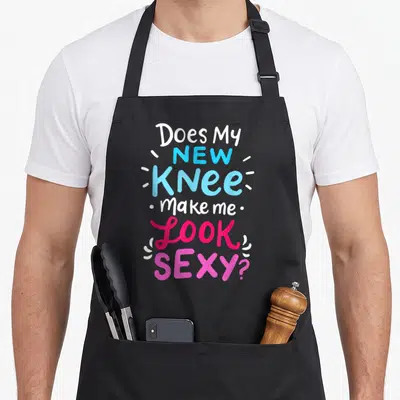 Knee joke apron - Stacked text Does My New Knee Make Me Look Sexy in white blue and pink — shown on a black cotton canvas apron against a white background