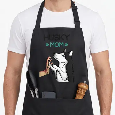 Mummy dog apron - Cartoon husky high-fiving human hand with HUSKY MOM text and teal paw prints — shown on a black cotton canvas apron against a white background